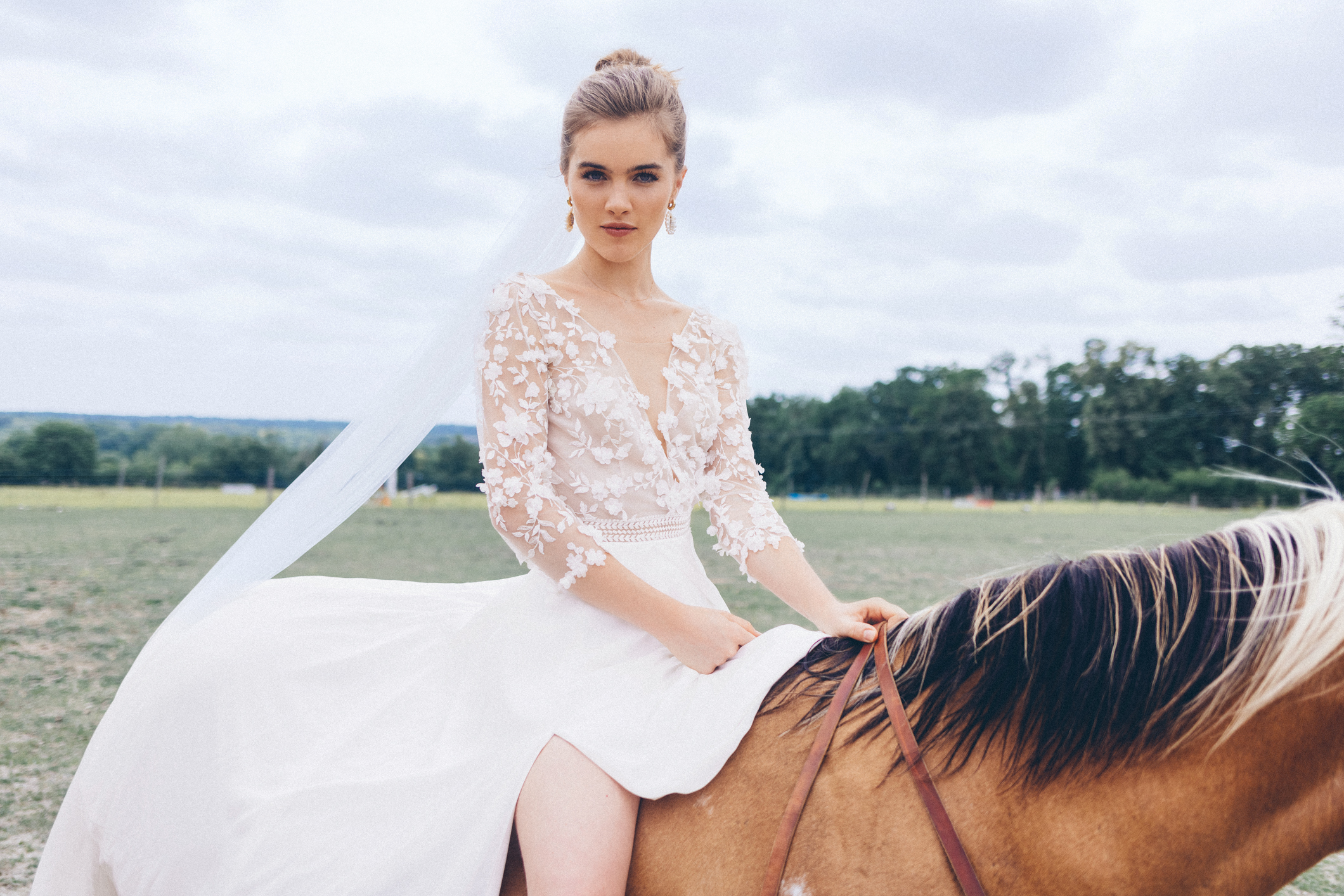 Mariée élégante en robe blanche en dentelle florale, posant à cheval dans un champ verdoyant sous un ciel nuageux — photo artistique pour boutique de robes de mariée.