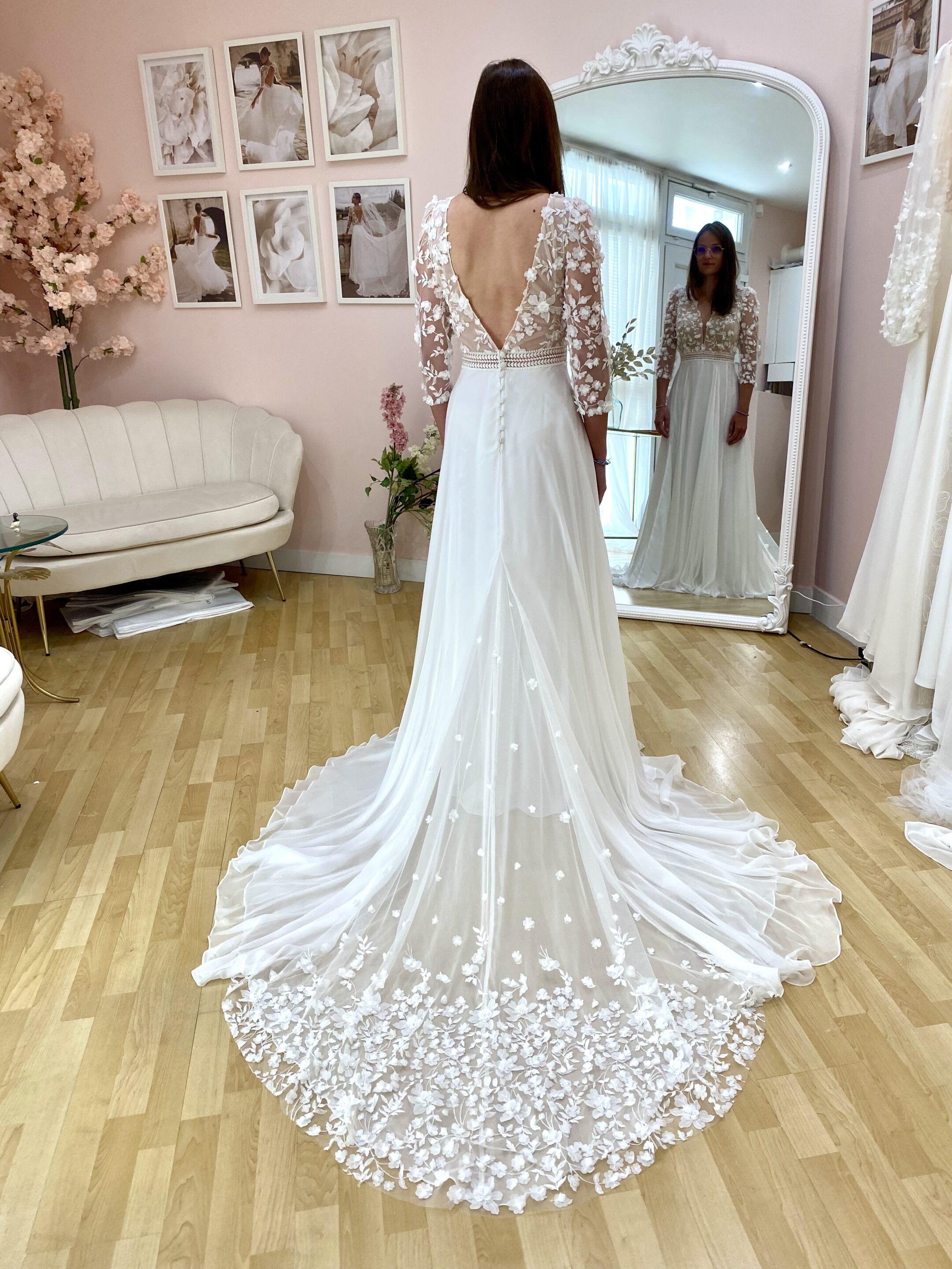 Robe de mariée Juliette avec manches longues transparentes ornées de dentelle florale, dos nu en V profond et jupe fluide prolongée par une traîne délicatement brodée. Essayage en boutique dans un décor raffiné, mettant en valeur le style romantique et sophistiqué de cette création.